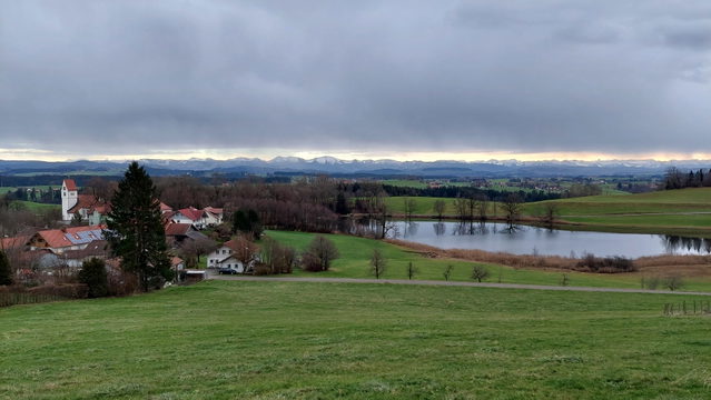 Landschaft mit kleinem Dorf, Kirche, Wiesen und See unter wolkigem Himmel.