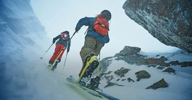Zwei Skifahrer klettern bergauf durch Schnee in felsiger Gebirgslandschaft.