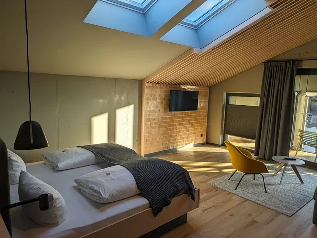 Modernes Hotelzimmer mit Doppelbett, gelbem Sessel, TV an Ziegelwand und Dachfenstern.