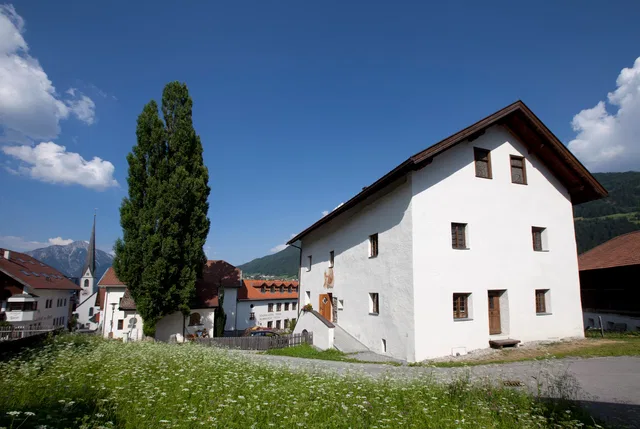 Weiße mehrstöckige Haus in einer ländlichen Ortschaft, neben einem hohen Baum, unter blauem Himmel.