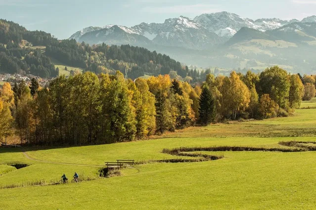 Zwei Radfahrende zwischen Wiesen und Herbstbäumen vor einer Bergkette