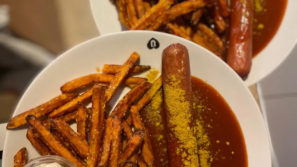 Zwei Teller Pommes frites mit einer langen Wurst in brauner Sauce, dazu Dipcreme mit Kräutern.
