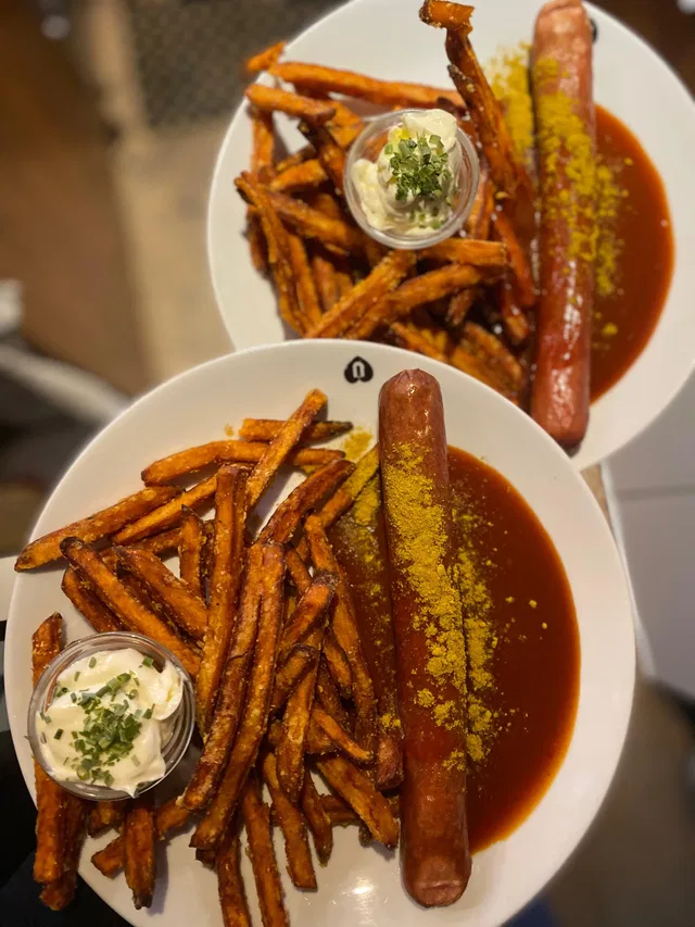 Zwei Teller Pommes frites mit einer langen Wurst in brauner Sauce, dazu Dipcreme mit Kräutern.