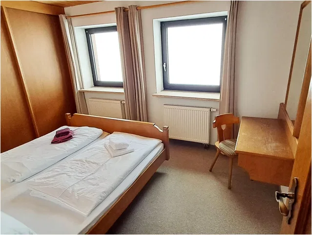 Kleines Hotelzimmer mit Doppelbett, zwei Fenstern, Schreibtisch und Stuhl.