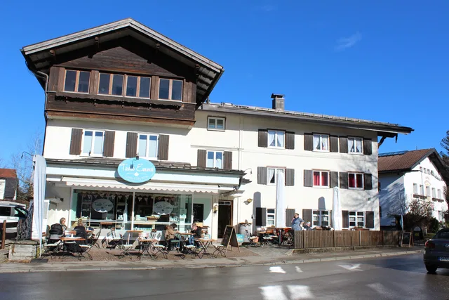 Straßencafé vor hellem Mehrfamilienhaus mit Holzbalkonen, sonniger Tag