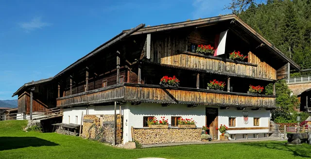 Traditionelles Bauernhaus mit Holzbalkonen und roten Blumen vor bewaldeten Bergen
