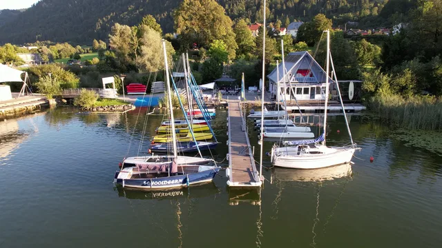 Boote am Steg eines ruhigen Sees, Uferhäuser und Wald im Hintergrund.