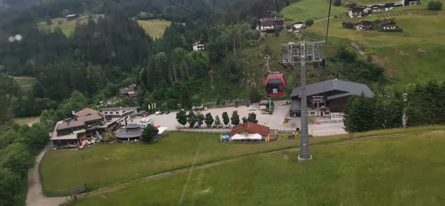 Bergdorf mit Seilbahnstationen, roter Gondel am Lift, grüne Wiesen und Wald im Hintergrund.
