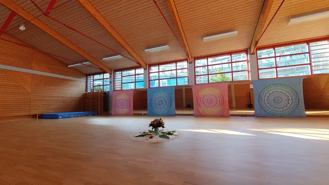 Heller Yogahallenraum mit Holzboden, großen Fenstern und bunten Mandala-Teppichen; in der Mitte liegt ein Blumengesteck.