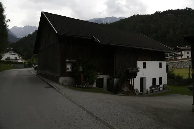 Große dunkle Scheune am Straßenrand mit angrenzendem weißen Gebäudeteil in einer Alpenlandschaft.