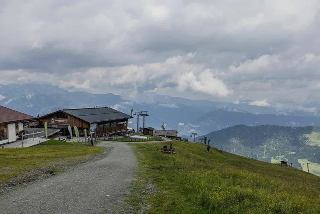 Bergrestaurant/Hütte am Almenweg mit Weg, Skianlagen im Hintergrund und Bergketten im Wolkenhimmel.