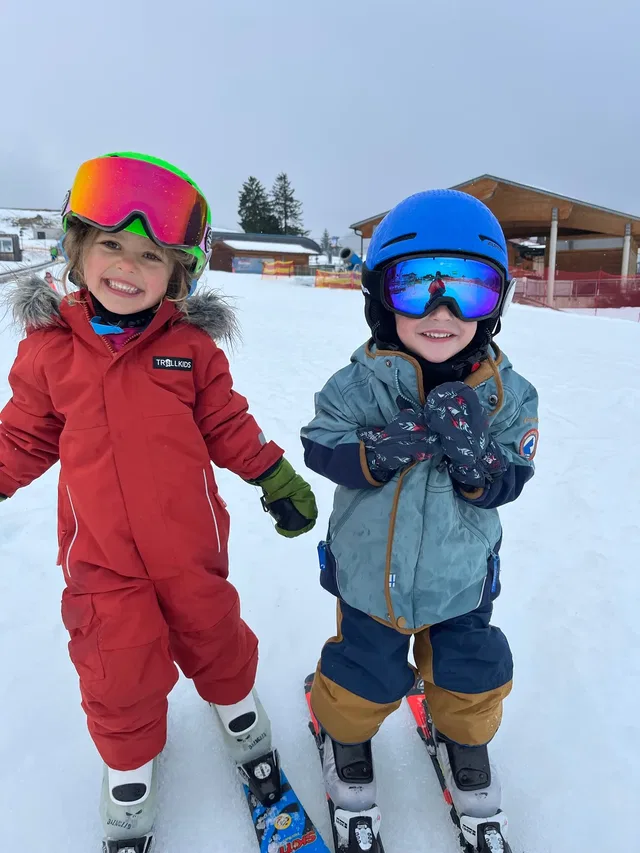 Zwei Kinder in Skianzügen mit Helmen stehen lachend im Schnee.