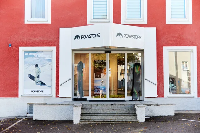 Frontalansicht des Powstore Kempten mit roter Fassade, Eingang und Snowboardpostern.