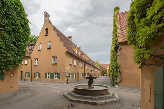 Steinbrunnen auf einem historischen Platz mit ockerfarbenen Häusern und grünen Fensterläden