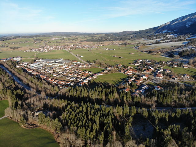 Luftaufnahme eines Dorfes mit Feldern, Wald und Bergen im Hintergrund.