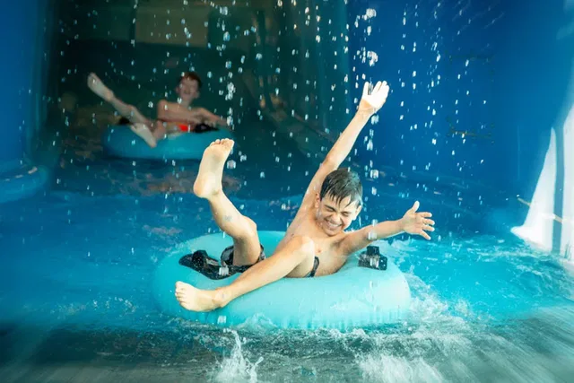 Zwei Jungen auf blauen Reifen in einer Wasserrutsche, der vordere hebt die Arme und lacht.