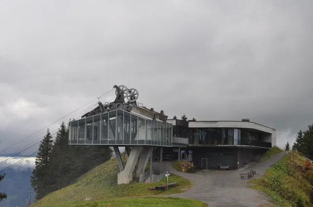 Moderne Glasarchitektur auf Bergkuppe mit Seilbahnradsammler-Dachstruktur und Aussicht auf Wald.