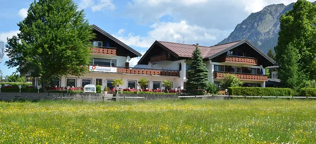 Fuggerhof-Gebäude mit Holzbalkonen, Terrasse und grüner Wiese vor Berglandschaft.