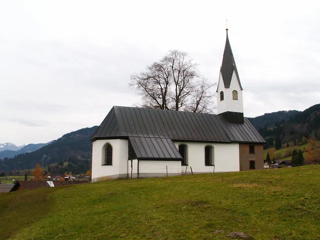 Kleine weiße Kirche mit dunklem Satteldach und spitzem Turm auf grünem Hügel vor Berglandschaft.