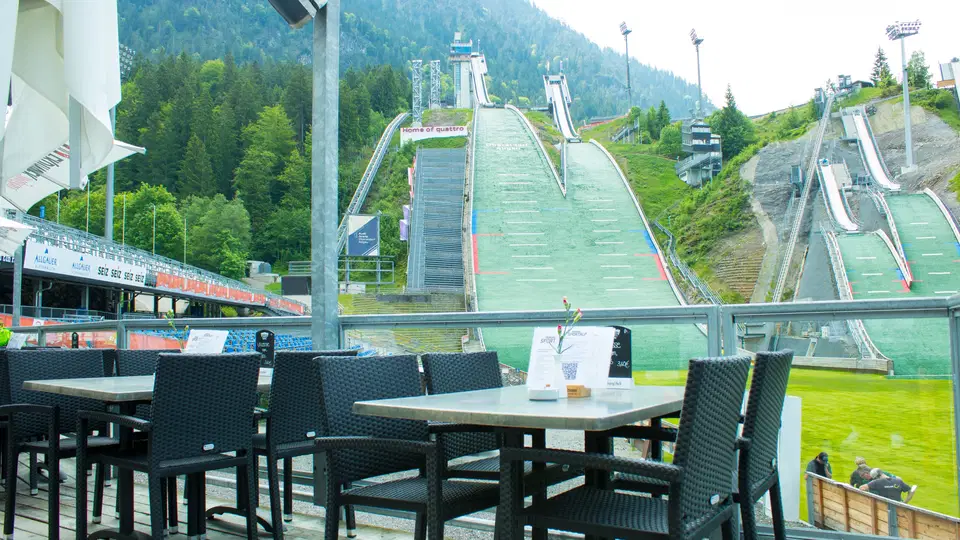 Outdoor-Café mit schwarzen Stühlen und Holztischen, Blick auf Skisprung-Schanzen im Hintergrund.