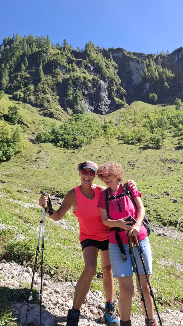 Zwei Wanderinnen mit Stöcken vor grünen Berghängen.