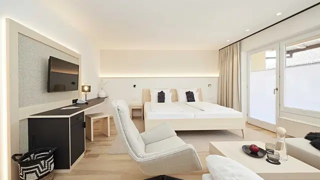 Helles Hotelzimmer mit Doppelbett, Lounge-Sessel, Schreibtisch und TV an der Wand.