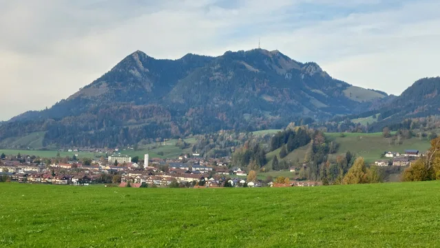 Landschaft mit grünem Feld im Vordergrund, kleinem Ort am Hang, Grünten-Massiv im Hintergrund.