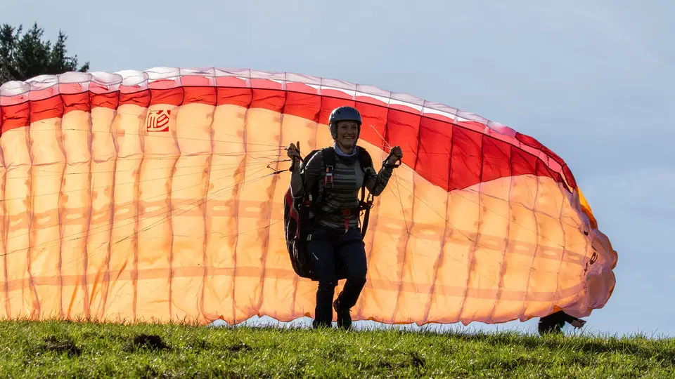 Person mit Helm und Gurtzeug beim Aufziehen eines orangefarbenen Gleitschirms am Übungshang.