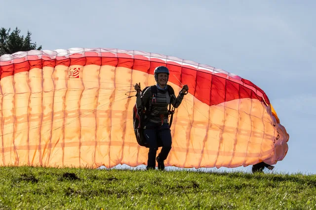 Person mit Gleitschirm-Ausrüstung auf grasiger Wiese vor orangefarbenem Paraglider