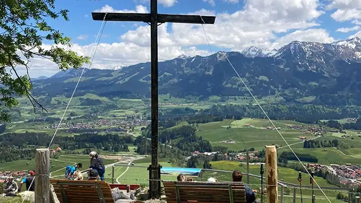 Großes Holzkreuz auf Aussichtspunkt mit Blick auf Alpenlandschaft und Dorf unten.