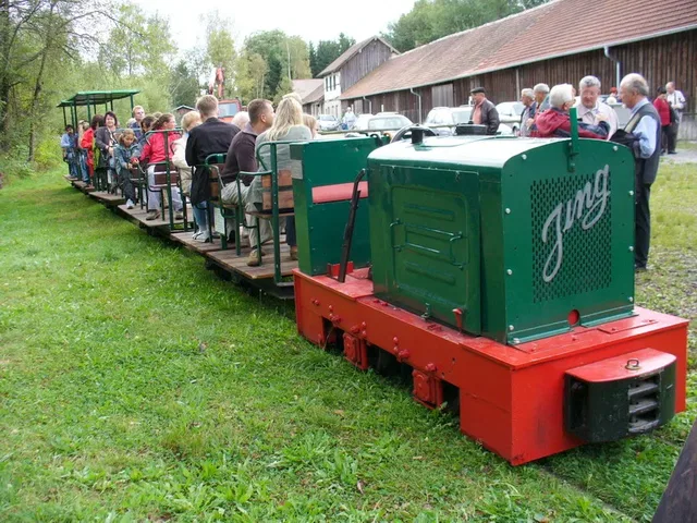 Kleiner grüner Mini-Zug mit rotem Unterbau und Passagieren am Bahnsteig