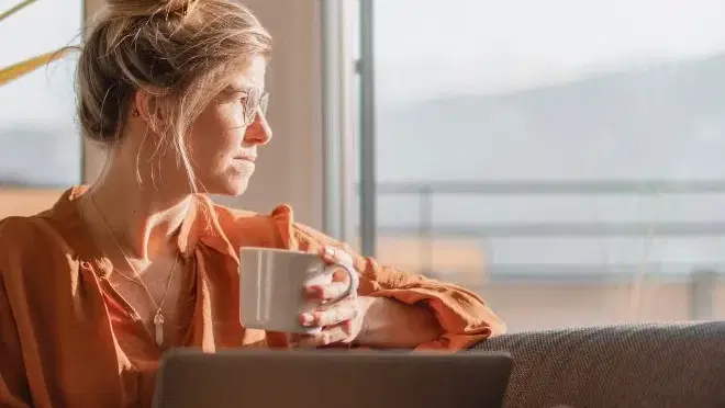 Frau sitzt auf Couch am Fenster, hält Tasse und arbeitet am Laptop