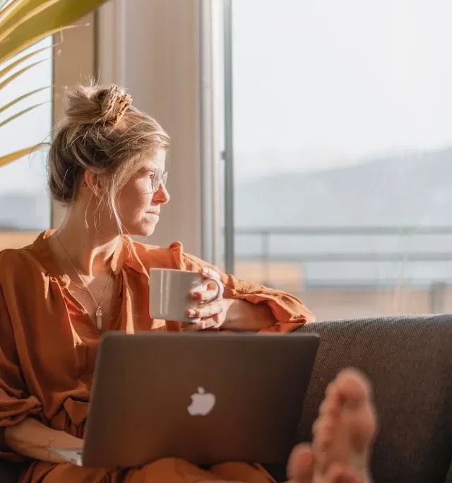 Frau sitzt auf Couch am Fenster, hält Tasse und arbeitet am Laptop