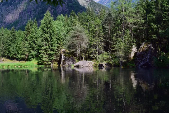 Spiegelnder See vor Wald und Felsen, Berge im Hintergrund.