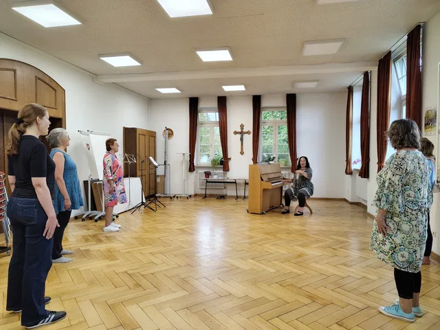 Gruppe von Erwachsenen steht im hellen Saal, links nebeneinander, vorne rechts Person am Klavier
