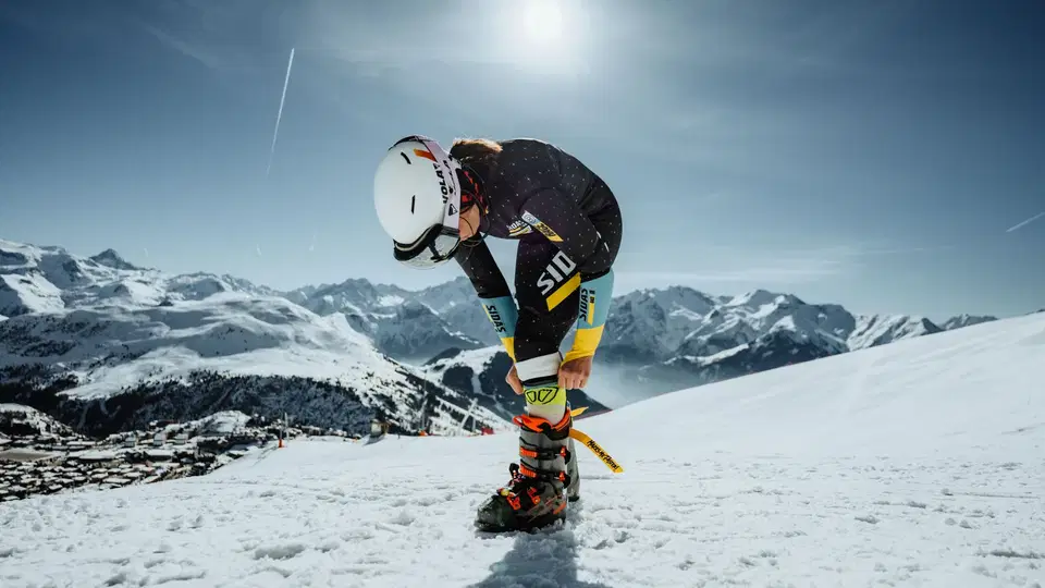 Skifahrer in Helm kniet auf Schnee und justiert Skibindung, Alpen im Hintergrund.