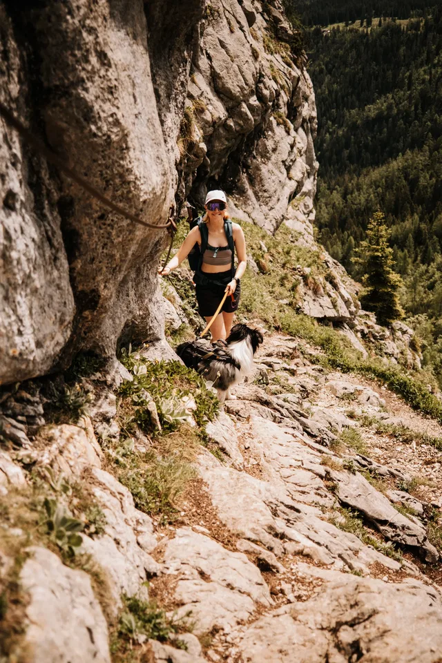 Wanderin mit Hund auf felsigem Bergpfad neben einer Felswand