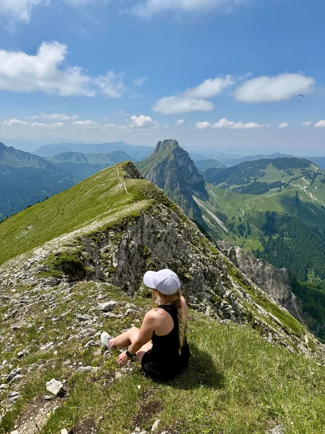 Frau mit weißer Kappe sitzt auf Bergkamm mit Blick auf Gipfel und Täler