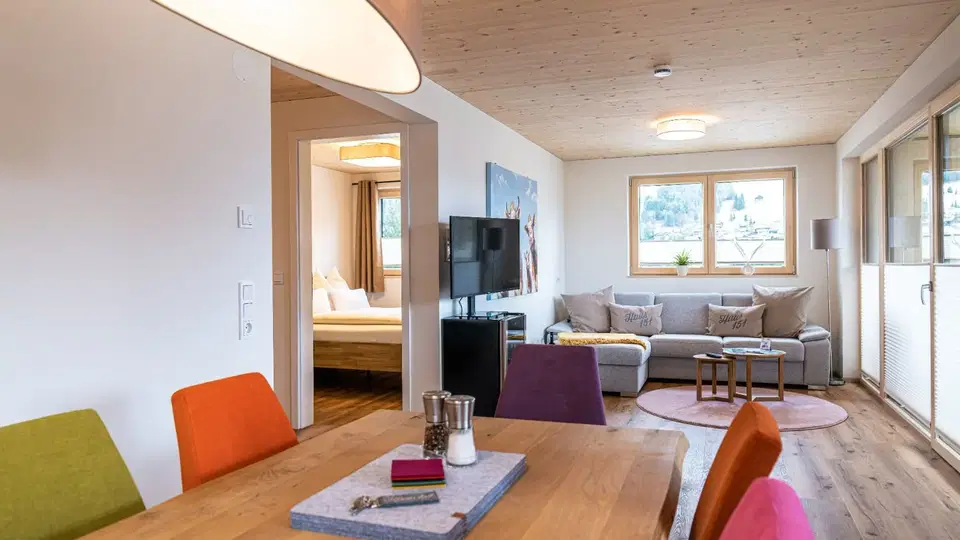 Offener Wohn- und Essbereich mit bunten Stühlen, Sofa und TV; Schlafzimmer durch Tür sichtbar.