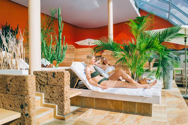 Zwei Personen entspannen auf Liegen in einem Spa mit Palmen und Kakteen.