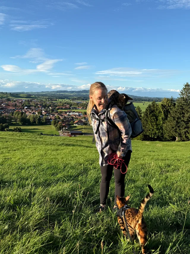 Frau mit Rucksack und Leine auf einer Grasdüne, Hund davor, Dorflandschaft im Hintergrund.