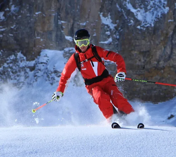 Skifahrer in roter Skiausrüstung fährt seitwärts durch Pulverschnee, Bergkulisse hinten
