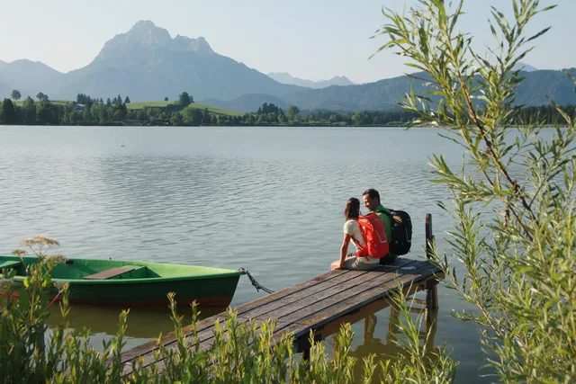 Paar mit Rucksack sitzt auf einem Holzsteg am See, Berge im Hintergrund