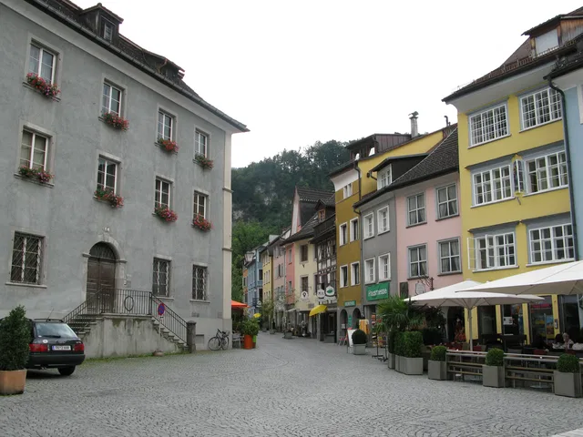 Bunte Häuserzeile am gepflasterten Platz mit Straßencafé und Fahrrad neben einer Steinfassade.