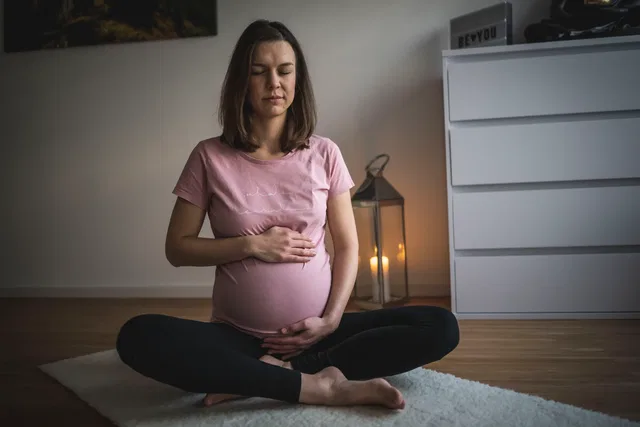 Schwangere Frau sitzt im Meditationssitz auf einem Teppich, Hände auf dem Bauch