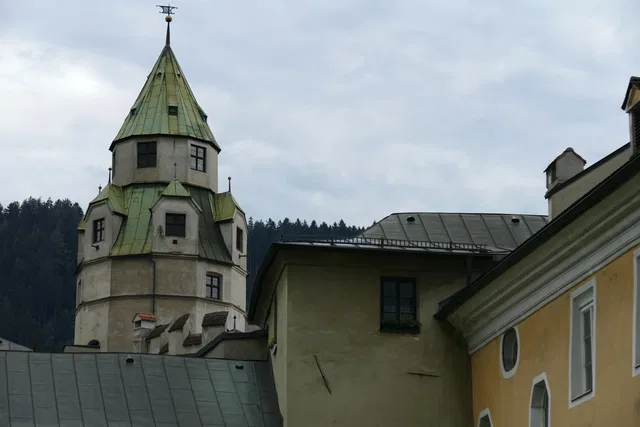 Historischer Gebäudekomplex mit grünem Turmdach vor einem bewaldeten Hang
