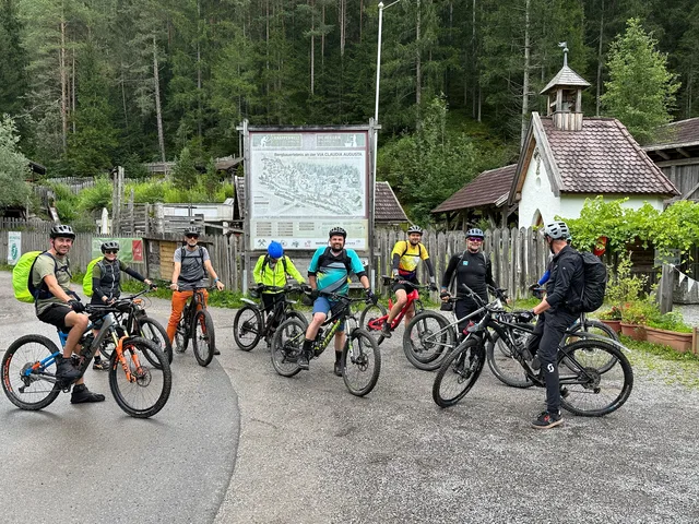 Sieben Mountainbiker vor historischen Holzbauten und einer Informationstafel im Wald