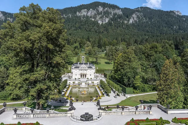 Barockgarten mit Treppen, Brunnenterrasse und weißem Schloss im Zentrum, umgeben von Wald und Bergen