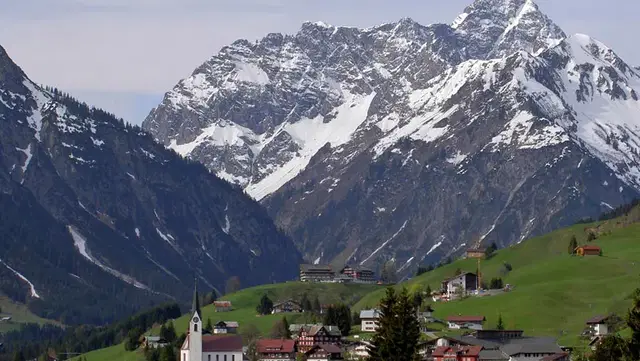 Ortsmitte von Hirschegg mit Kirche, grünen Wiesen und schneebedeckten Berggipfeln im Hintergrund.