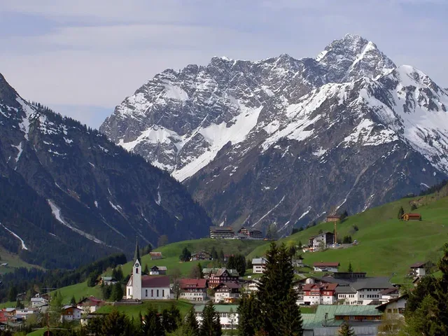 Bergdorf mit Kirche, grüne Wiesen und schneebedeckte Gipfel im Hintergrund